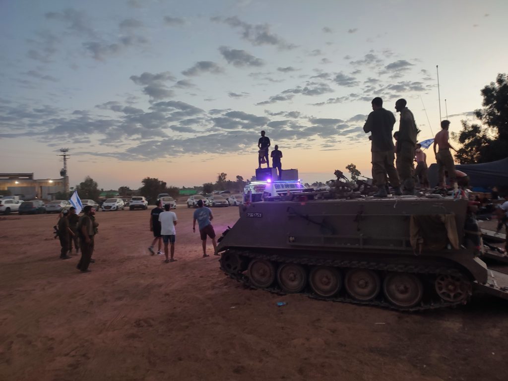 חיילים ואזרחים חוגגים ליד נגמ"ש עם רכב נחמנים משמחים, שקיעה מרהיבה ברקע